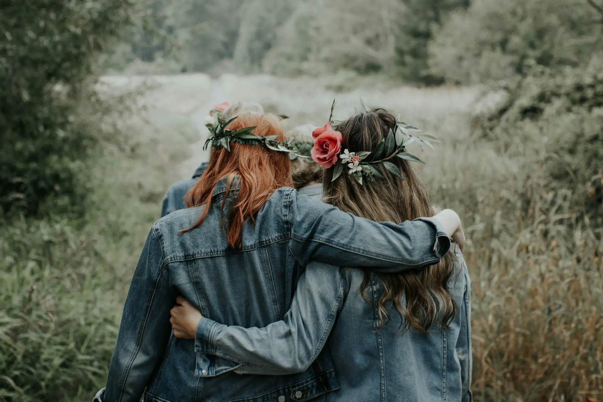 view of two women from behind with their arms around each other, wearing jean jackets and pink and green flower crowns