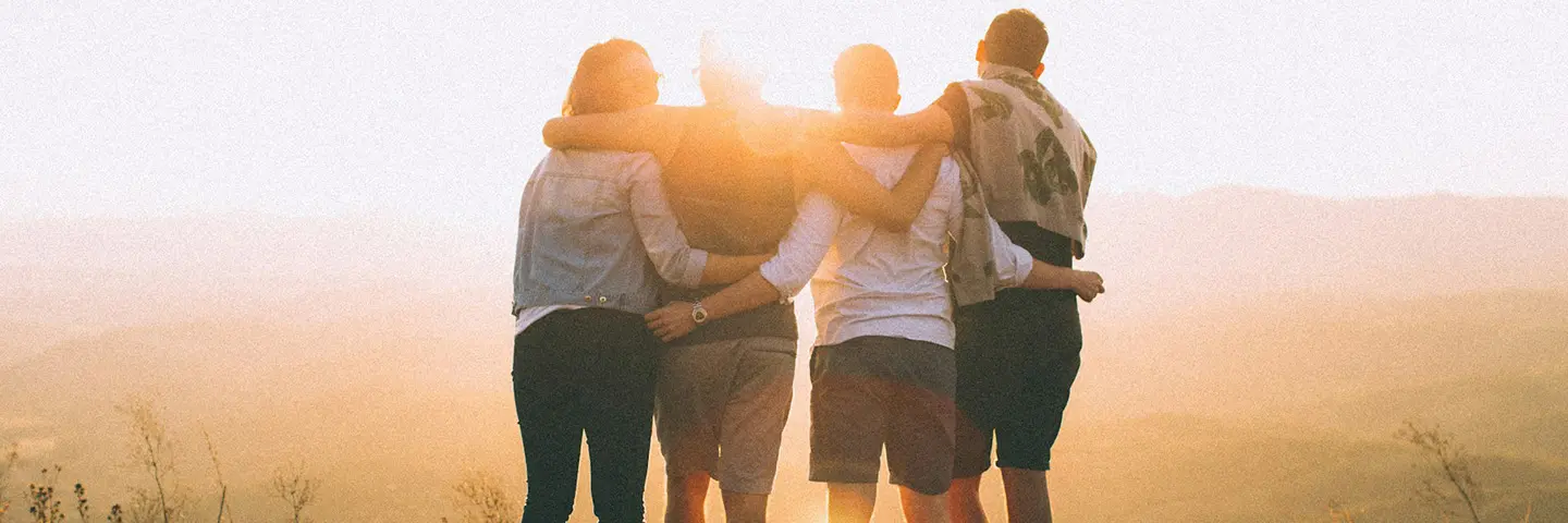 view from behind of four friends with their arms around each other staring out at a sunset over a mountain